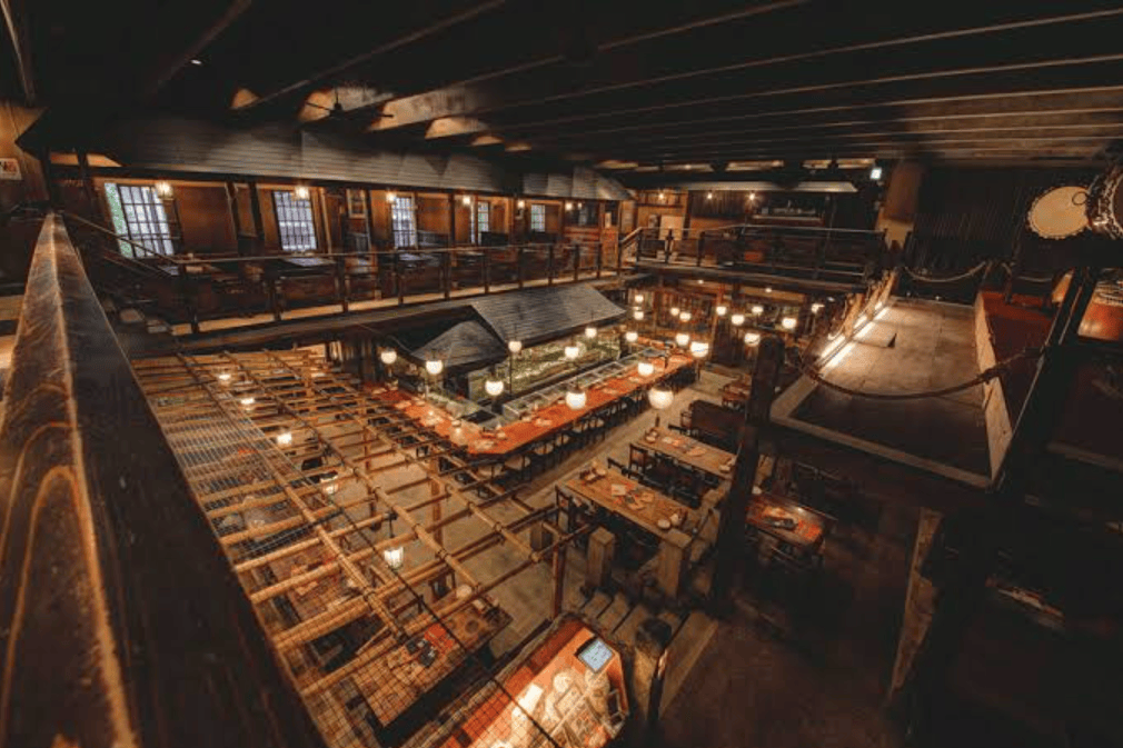 An aerial view of a spacious restaurant interior featuring wooden beams, multiple dining areas, and warm lighting. Tables are arranged across different levels, with a central serving area visible.