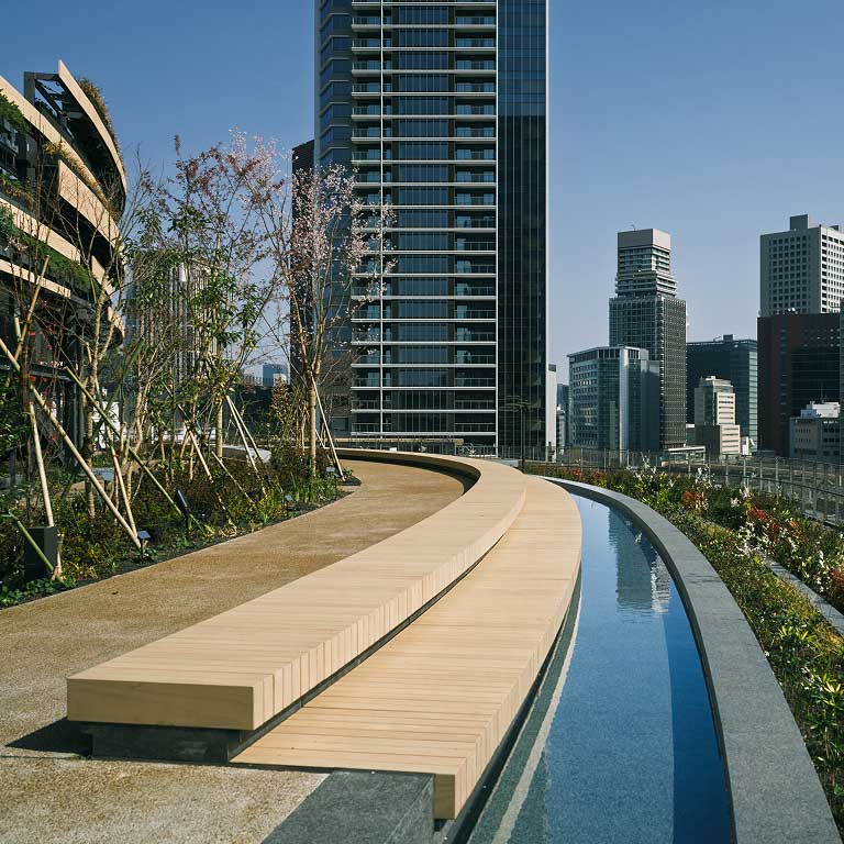 Modern urban rooftop landscape featuring a curved wooden bench, a narrow water feature, and city skyline in the background.